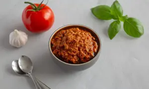 A miseczce znajduje się aromatyczna pasta z suszonych pomidorów, obok świeży pomidor, czosnek i listki bazylii.