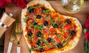 Serce z pizzy z rukolą i oliwkami, idealne na romantyczną kolację w domu. Obok złote sztućce i młynek do pieprzu.
