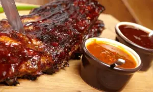 Żeberka w sosie BBQ, idealne do domowego sosu bbq przepis. Nóż wbity w mięso, obok miseczki z sosem.