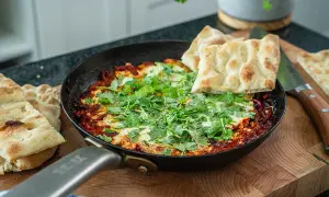 Szakszuka co to? Aromatyczne jajka w sosie pomidorowym z kolendrą, podane z chlebkiem pita na desce.