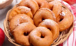Puszyste jabłka w cieście, posypane cukrem pudrem, kuszą w wiklinowym koszyku. Obok czerwone jabłka i miseczka mąki.