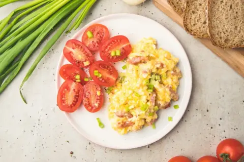 Pyszna kolacja z jajek z pomidorkami i szczypiorkiem. Obok kromki chleba i pęczek zielonej cebulki.