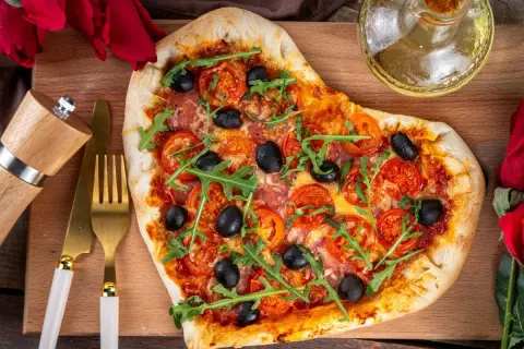 Serce z pizzy z rukolą i oliwkami, idealne na romantyczną kolację w domu. Obok złote sztućce i młynek do pieprzu.