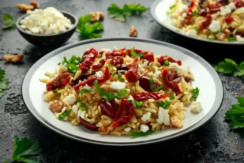 Pyszna sałatka z makaronem orzo, suszonymi pomidorami, fetą i oliwkami, udekorowana natką pietruszki.