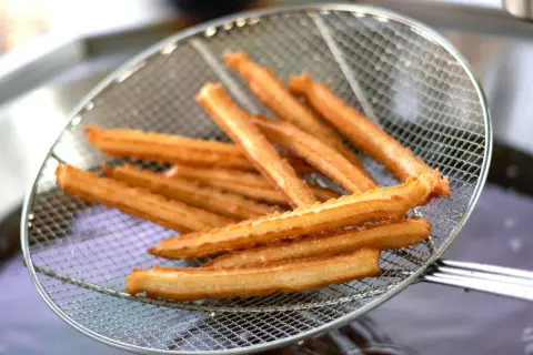 Churros przepis: złociste, chrupiące paluszki wyjęte z frytownicy na metalowej łyżce cedzakowej.