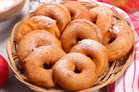Puszyste jabłka w cieście, posypane cukrem pudrem, kuszą w wiklinowym koszyku. Obok czerwone jabłka i miseczka mąki.
