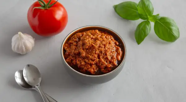 A miseczce znajduje się aromatyczna pasta z suszonych pomidorów, obok świeży pomidor, czosnek i listki bazylii.