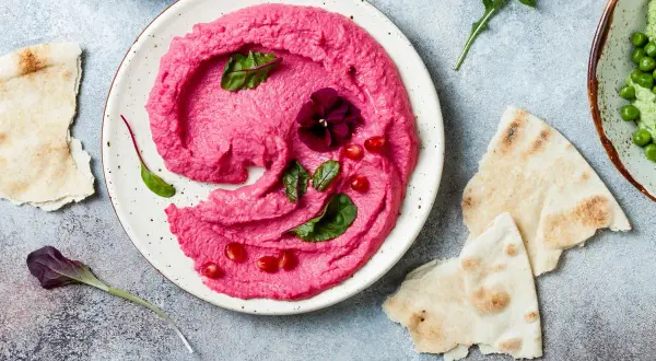Piękny, różowy hummus z buraka, udekorowany kwiatkiem i granatem, podany z pitą.