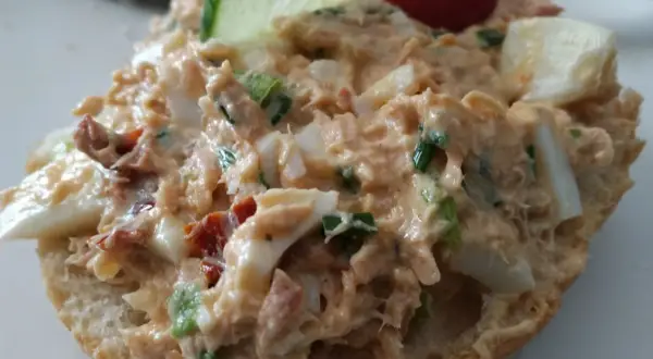 Pyszna pasta z tuńczyka z jajkiem i szczypiorkiem na bułce, ozdobiona plasterkiem ogórka i pomidorkiem.