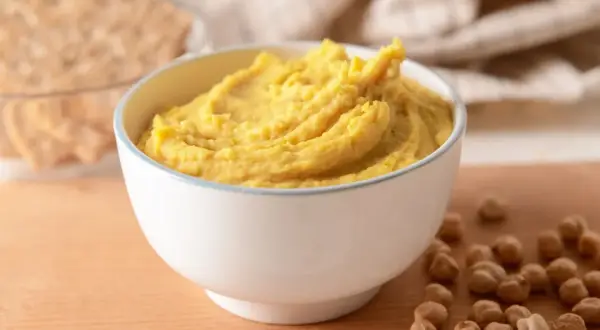 Puszysta pasta z ciecierzycy w miseczce, obok kilka ziaren ciecierzycy i chrupki.