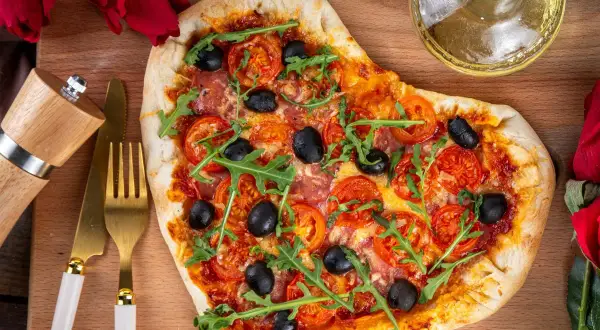Serce z pizzy z rukolą i oliwkami, idealne na romantyczną kolację w domu. Obok złote sztućce i młynek do pieprzu.