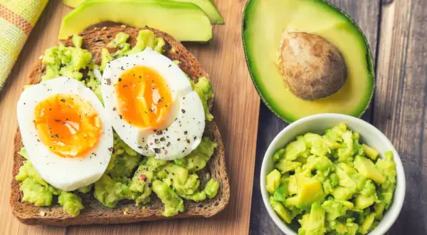 Pyszne awokado jak jeść: tost z rozgniecionym awokado i jajkiem na miękko, obok połówka awokado i miseczka z kawałkami awokado.
