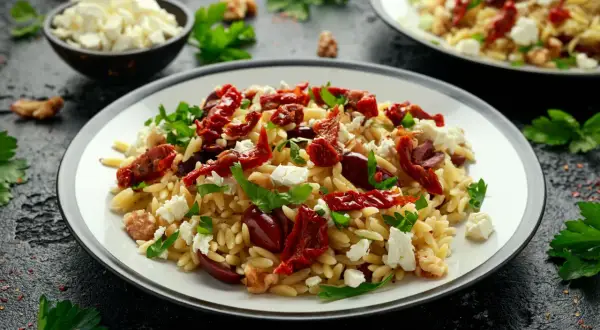 Pyszna sałatka z makaronem orzo, suszonymi pomidorami, fetą i oliwkami, udekorowana natką pietruszki.
