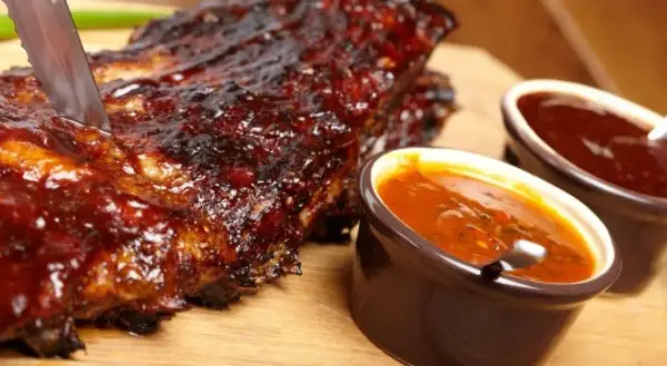 Żeberka w sosie BBQ, idealne do domowego sosu bbq przepis. Nóż wbity w mięso, obok miseczki z sosem.