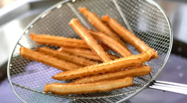 Churros przepis: złociste, chrupiące paluszki wyjęte z frytownicy na metalowej łyżce cedzakowej.