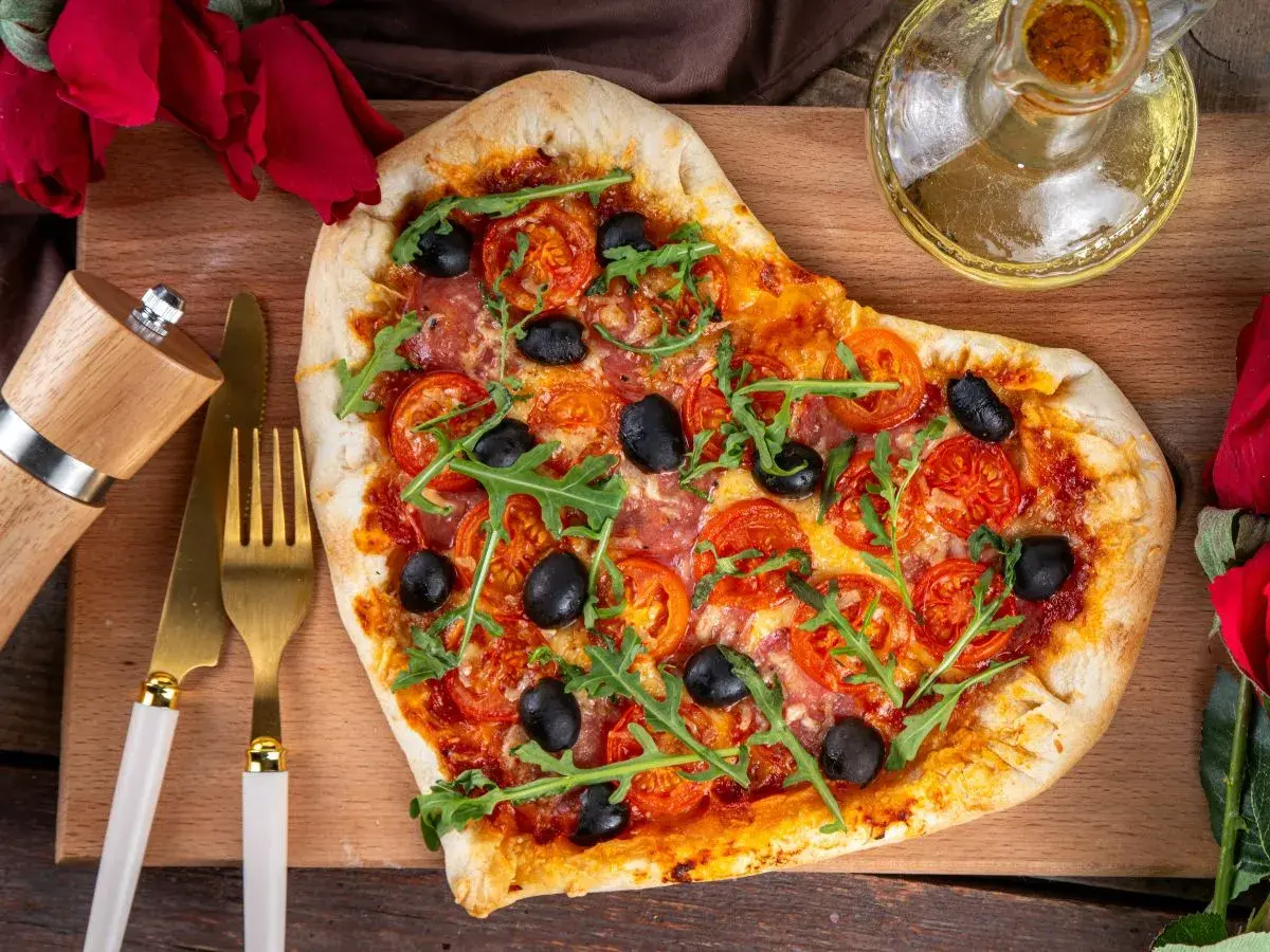 Serce z pizzy z rukolą i oliwkami, idealne na romantyczną kolację w domu. Obok złote sztućce i młynek do pieprzu.