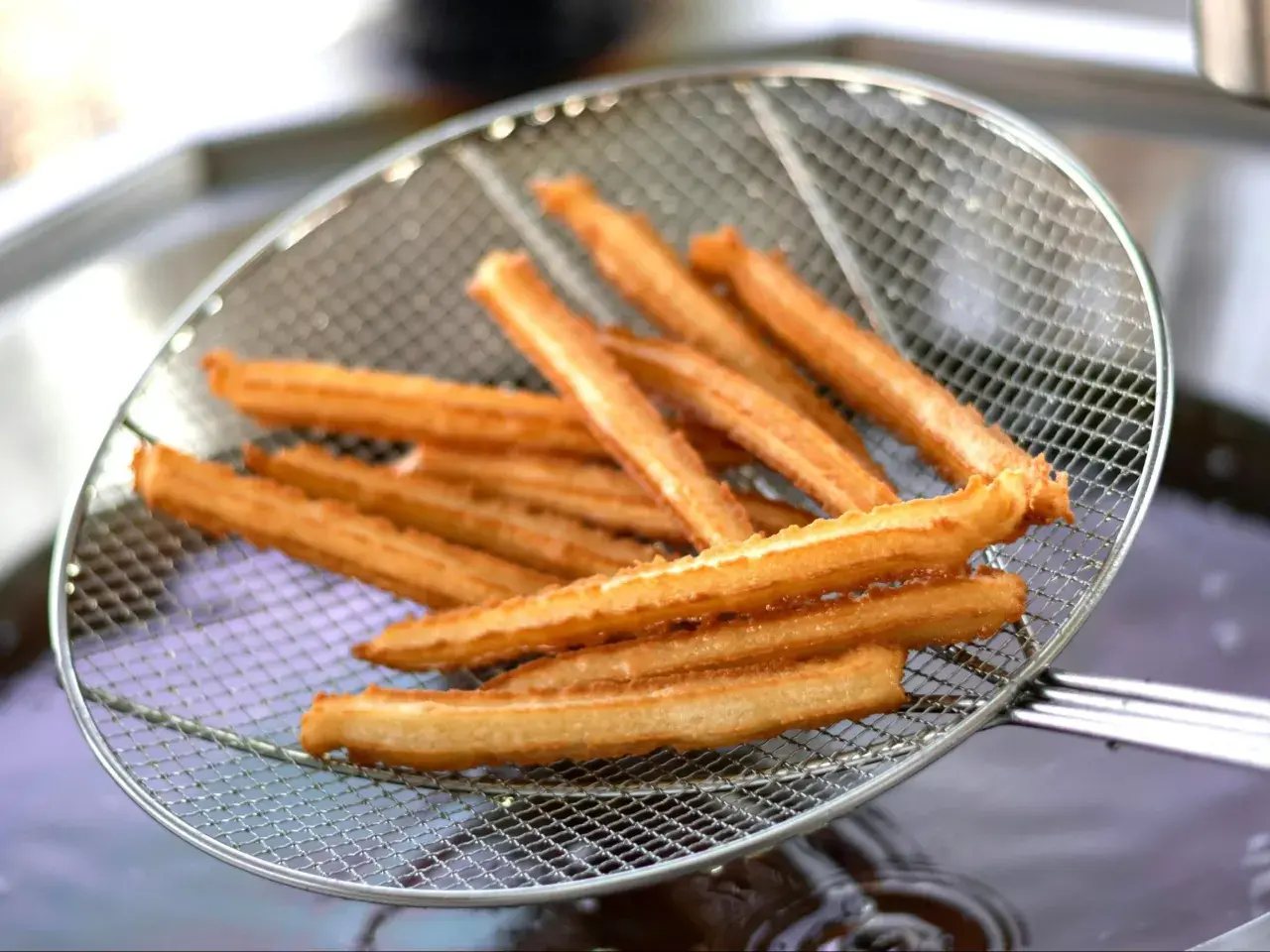 Churros przepis: złociste, chrupiące paluszki wyjęte z frytownicy na metalowej łyżce cedzakowej.