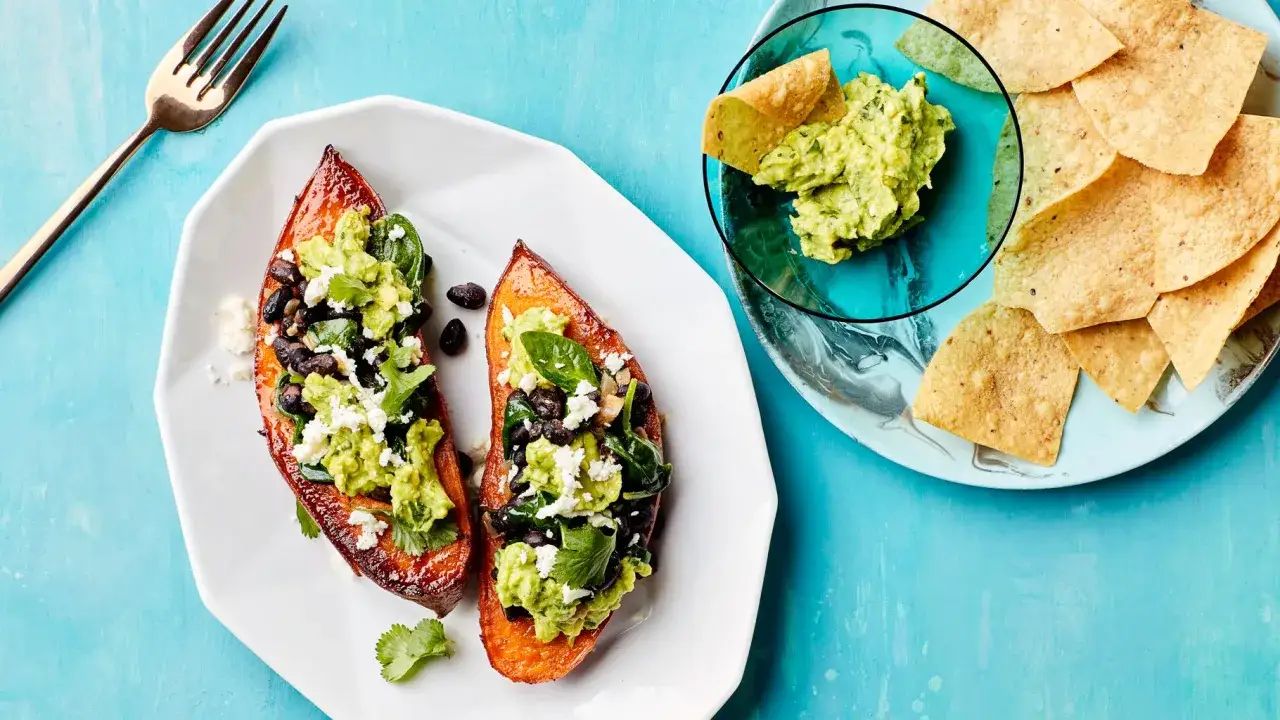 Dwa poł&oacute;wki pieczonych batat&oacute;w z czarną fasolą, awokado i serem feta. Obok miseczka guacamole z nachos.