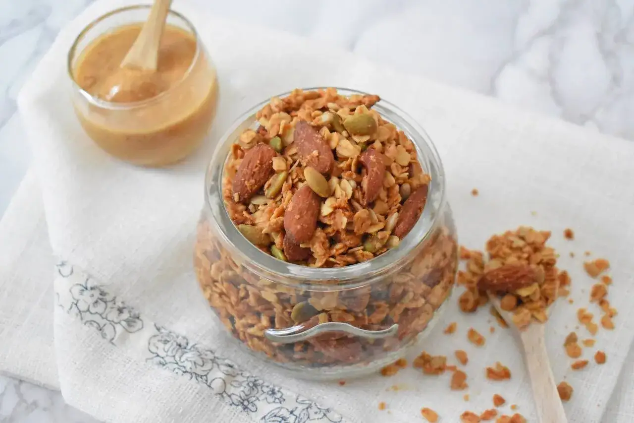 Domowy granola przepis pełen migdał&oacute;w i pestek dyni. Idealny na śniadanie, a obok słoiczek z masłem orzechowym.