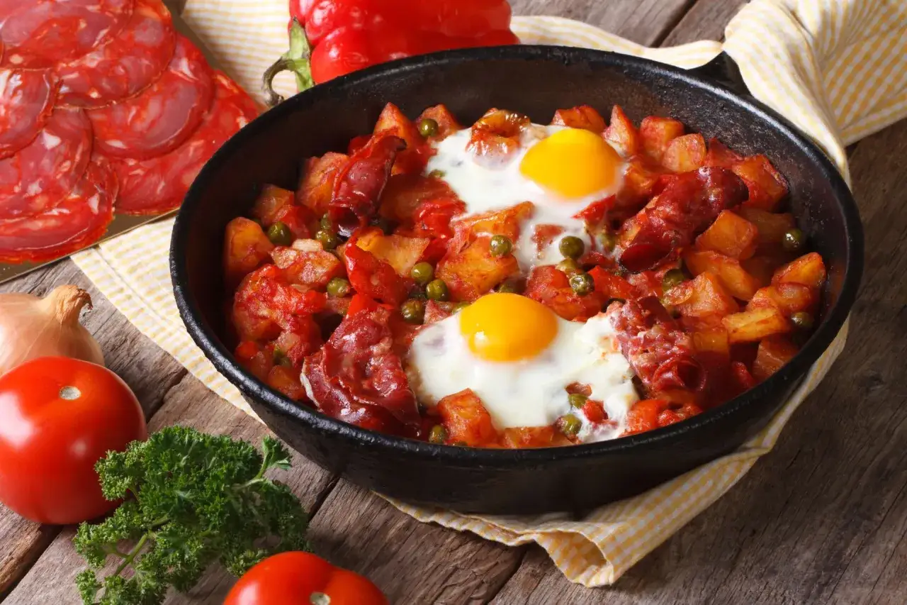 Pyszna kolacja z jajek, ziemniak&oacute;w, chorizo i groszku w patelni. Obok pomidory, cebula i natka pietruszki.