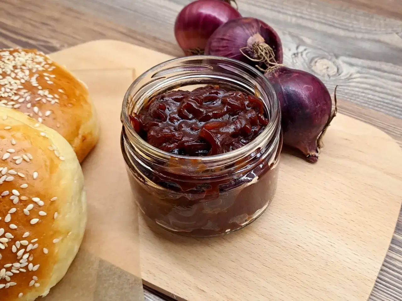 Słoiczek z ciemnym, błyszczącym sosem cebulowym, obok świeżych cebul i bułeczek do burger&oacute;w. Idealny dodatek do burger&oacute;w, sos cebulowy podkreśli smak.