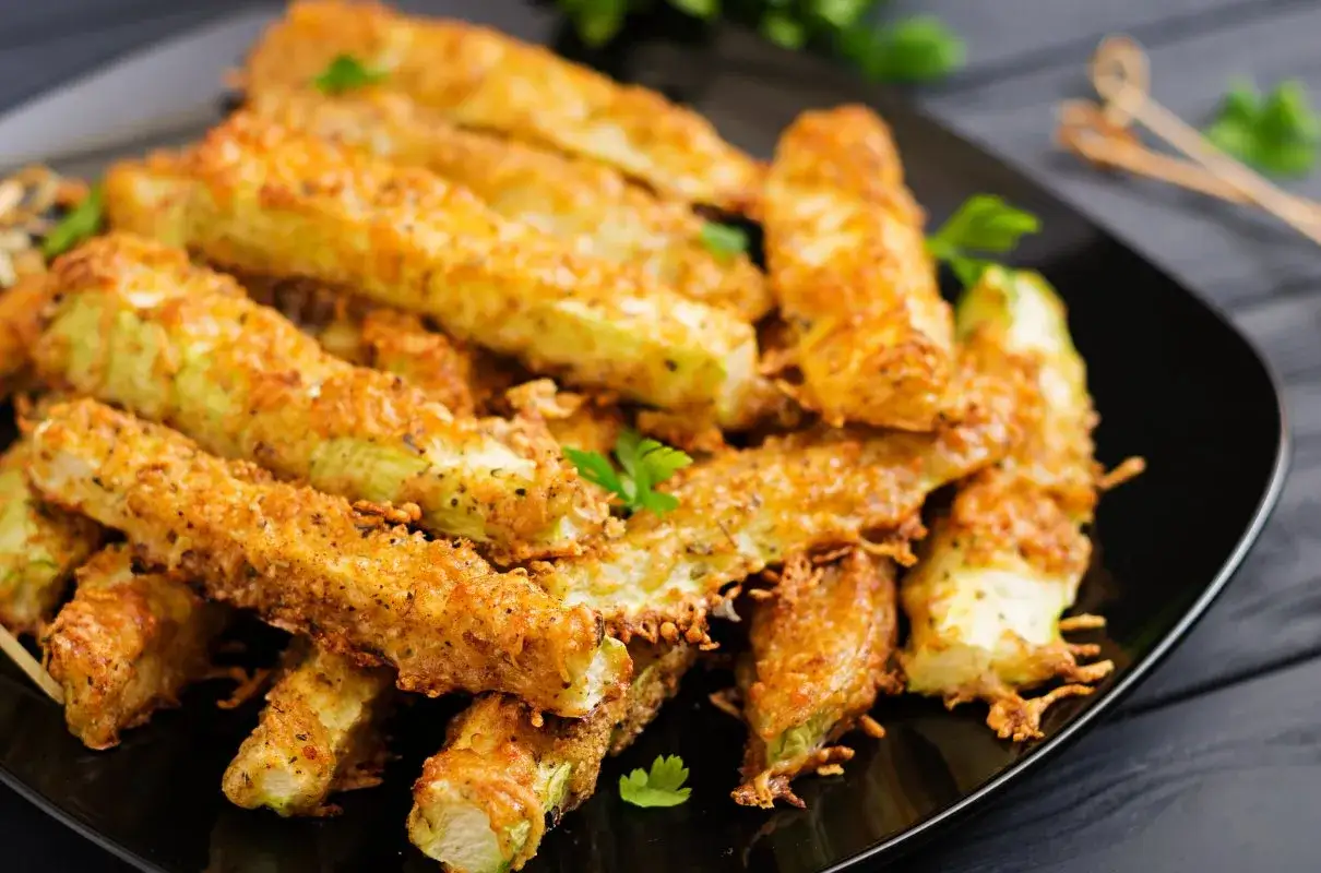 Chrupiące paluszki z cukinii w cieście, posypane parmezanem i ziołami, ułożone na czarnym talerzu.