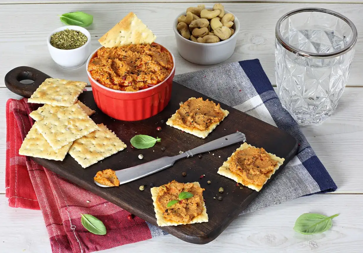 Pyszna pasta z suszonych pomidor&oacute;w na krakersach, gotowa do degustacji. Obok miseczka z orzechami i szklanka wody.