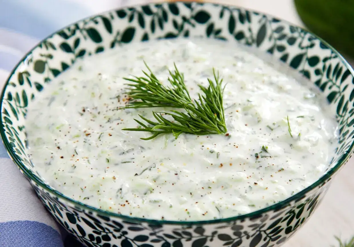 Kremowy sos tzatziki z kawałkami ogórka i koperkiem, doprawiony pieprzem, w ozdobnej misce.
