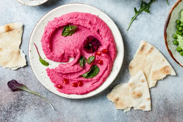 Piękny, różowy hummus z buraka, udekorowany kwiatkiem i granatem, podany z pitą.