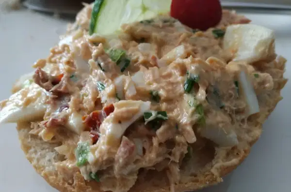 Pyszna pasta z tuńczyka z jajkiem i szczypiorkiem na bułce, ozdobiona plasterkiem ogórka i pomidorkiem.