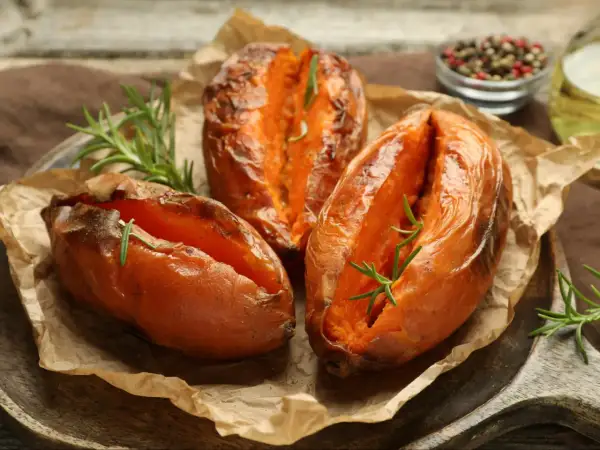 Trzy pieczone bataty z rozmarynem na papierze do pieczenia, wyglądają apetycznie i aromatycznie.