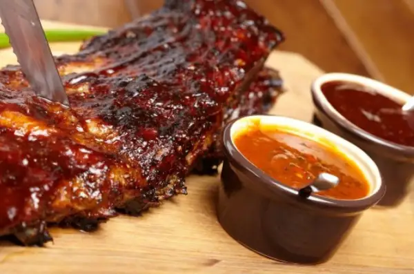 Żeberka w sosie BBQ, idealne do domowego sosu bbq przepis. Nóż wbity w mięso, obok miseczki z sosem.