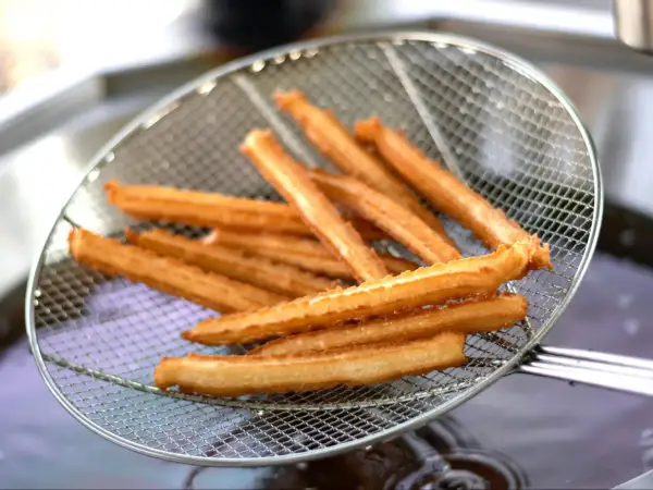 Churros przepis: złociste, chrupiące paluszki wyjęte z frytownicy na metalowej łyżce cedzakowej.
